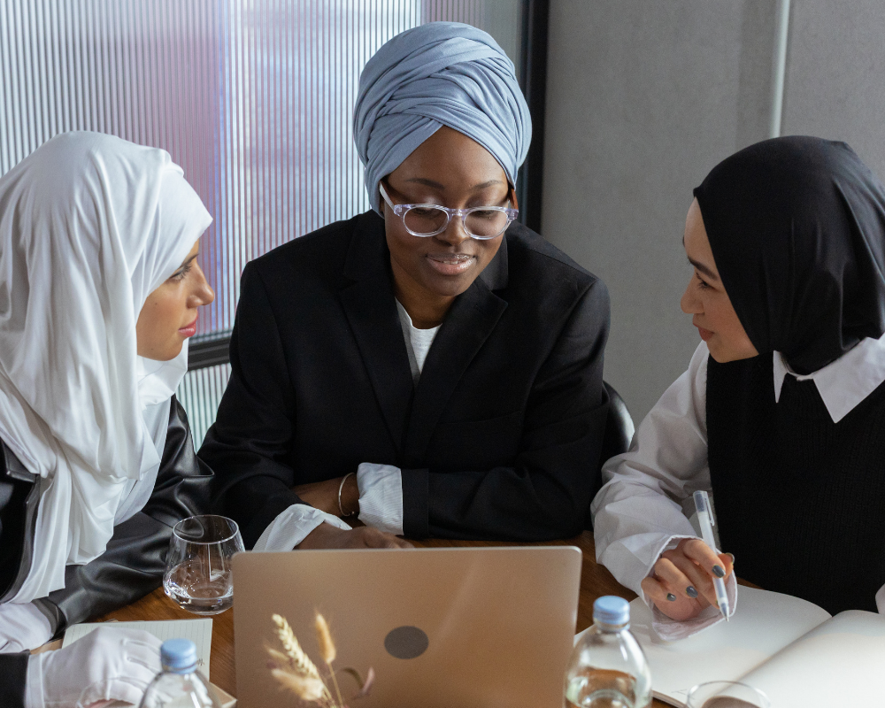Three women wearing hijabs sitting at a desk with a laptop, paper and various items on a table.