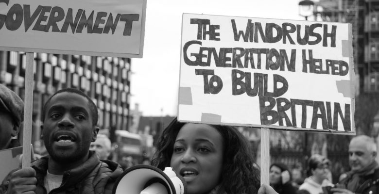 A black and white photo of people at a protest. A woman with a megaphone holds a placard saying "The Windrush Generation Helped to Build Britain" whilst a man stands to her left with another sigh.